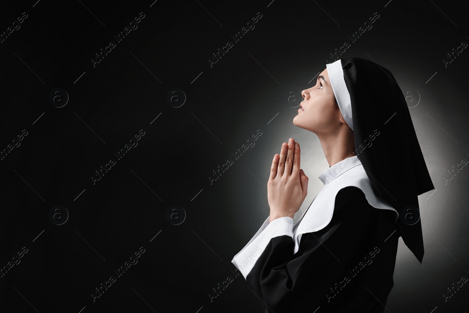 Photo of Nun with clasped hands praying to God on black background. Space for text