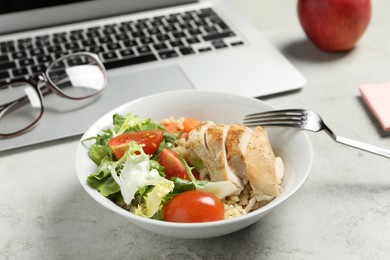 Photo of Bowl with tasty food, laptop, fork and glasses on light grey table, closeup. Business lunch