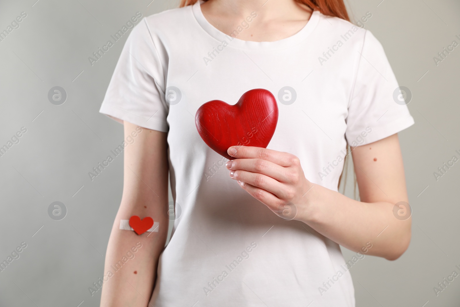 Photo of Blood donation concept. Woman with adhesive plaster on arm holding red heart against grey background, closeup