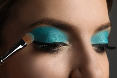 Photo of Applying cyan eye shadow onto woman's face on grey background, closeup. Beautiful evening makeup