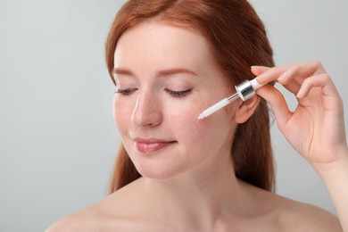 Photo of Beautiful woman with freckles applying cosmetic serum onto her face against grey background, closeup