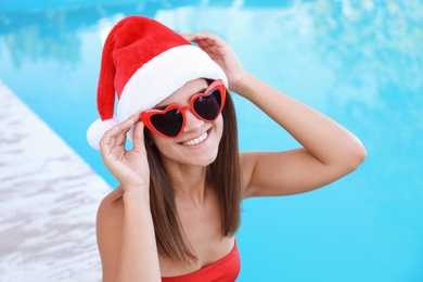 Photo of Young woman wearing Santa Claus hat near swimming pool. Christmas vacation