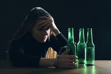 Alcohol addiction. Woman handcuffed to bottle of beer at wooden table against black background