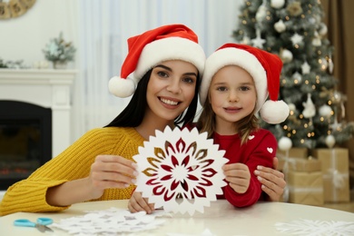 Photo of Mother and daughter in Santa hats with paper snowflake near Christmas tree at home