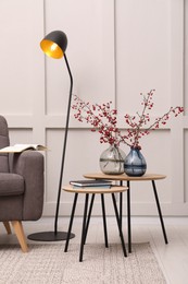 Photo of Hawthorn branches with red berries in vases, armchair and lamp indoors. Interior design