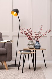 Hawthorn branches with red berries in vases, armchair and lamp indoors. Interior design