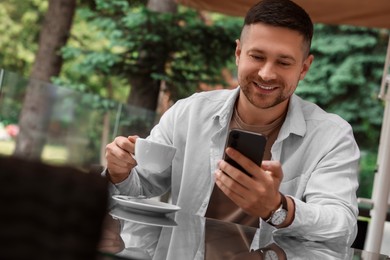 Photo of Handsome man with cup of drink sending message via smartphone at table in outdoor cafe