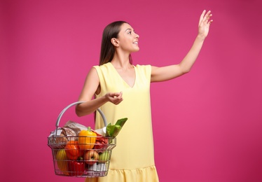 Young woman with shopping basket full of products on pink background