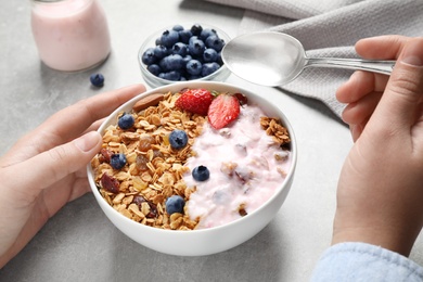 Woman eating tasty homemade granola with yogurt and berries at grey table, closeup. Healthy breakfast