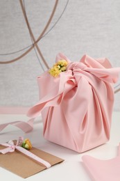 Furoshiki technique. Gift packed in pink fabric, card and flowers on white table
