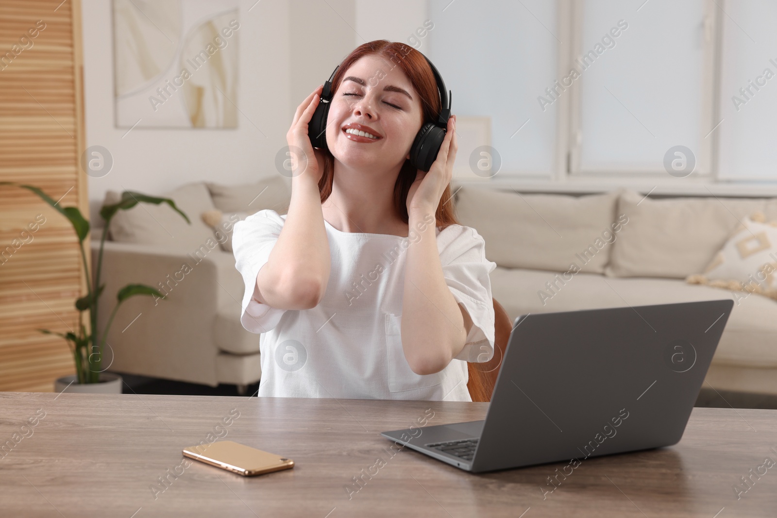 Photo of Happy woman with headphones listening to music near laptop at wooden table in room