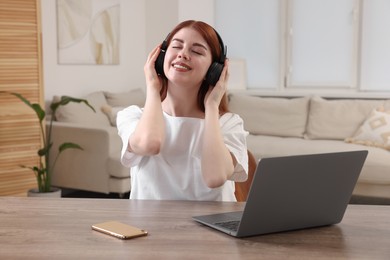 Happy woman with headphones listening to music near laptop at wooden table in room