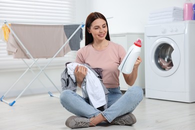 Woman sitting on floor near washing machine and holding fabric softener with dirty clothes in bathroom