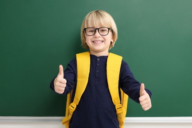 Photo of Happy little school child with backpack showing thumbs up near chalkboard