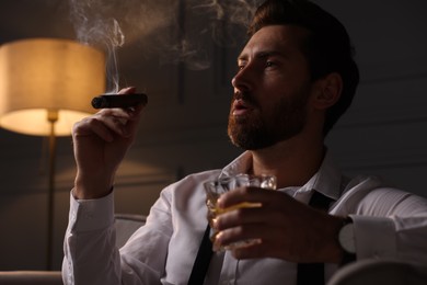 Photo of Handsome man with glass of whiskey smoking cigar at home