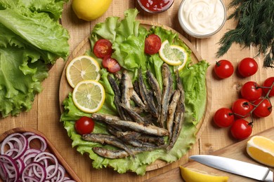 Photo of Delicious fried anchovies served with lemon, tomatoes and sauce on wooden table, flat lay