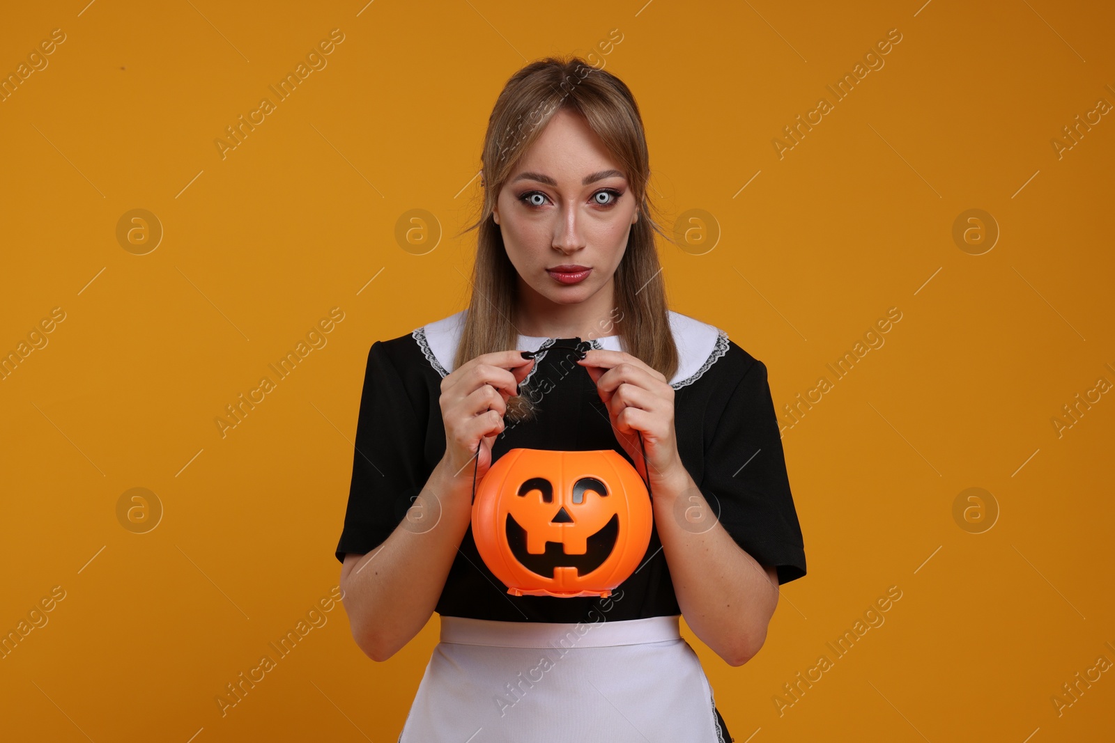 Photo of Woman in scary maid costume with pumpkin bucket on orange background. Halloween celebration