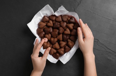 Woman with box of heart shaped chocolate candies at black table, top view