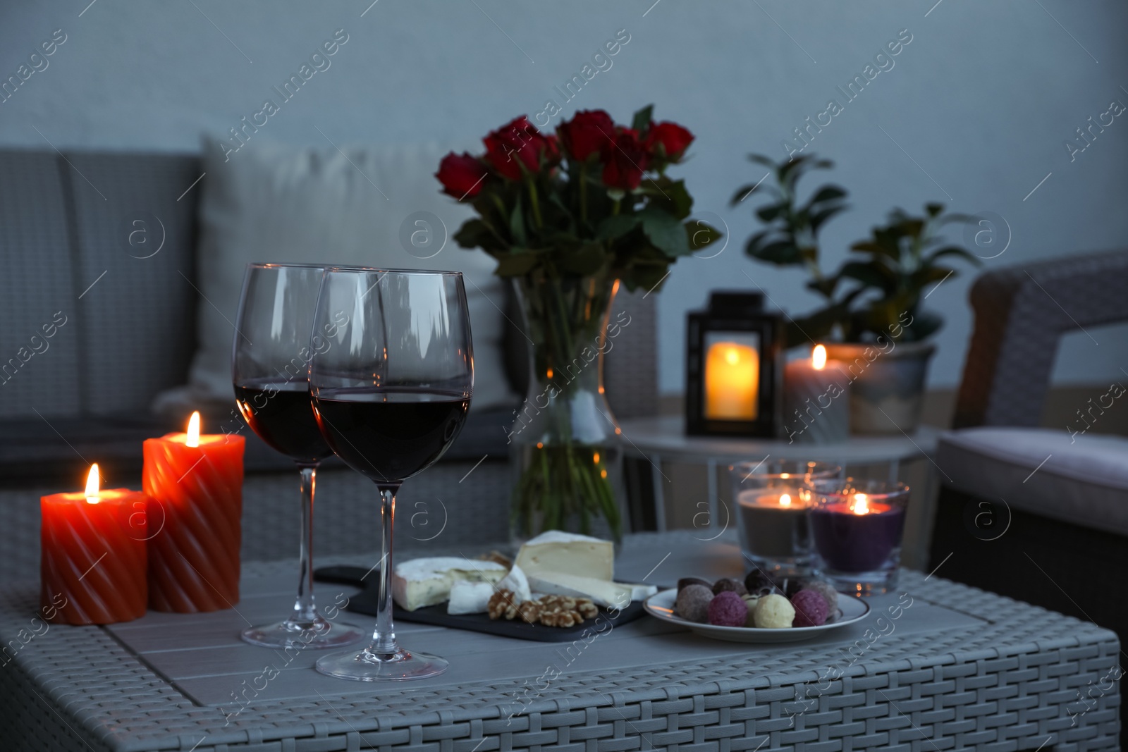 Photo of Glasses of wine, vase with roses, burning candles and snacks on rattan table at balcony in evening, closeup