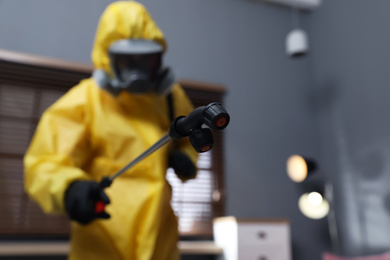 Pest control worker in protective suit indoors, focus on insecticide sprayer