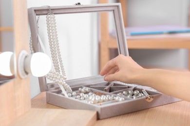 Photo of Young woman taking elegant pearl jewelry at dressing table indoors, closeup