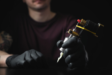 Tattoo artist with professional machine at table against black background, closeup