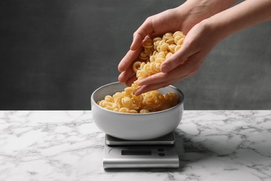 Photo of Woman adding pasta into bowl on kitchen scale on white marble table, closeup