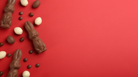 Photo of Flat lay composition with chocolate Easter bunnies, eggs and candies on red background. Space for text