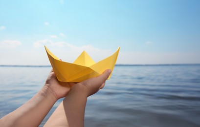 Child holding yellow paper boat near river, closeup. Space for text