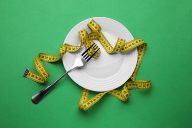 Plate, measuring tape and fork on green background, top view. Diet concept