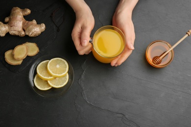 Photo of Woman holding cup of immunity boosting drink at dark table with ingredients, top view. Space for text