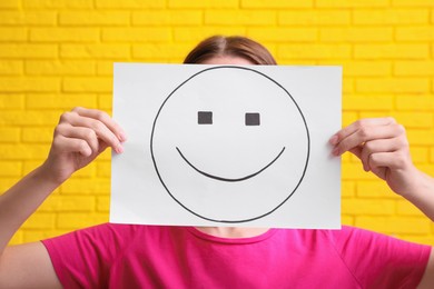 Woman hiding behind sheet of paper with happy face against yellow brick wall