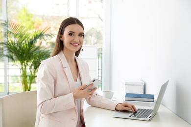 Photo of Young businesswoman with smartphone using laptop at table in office