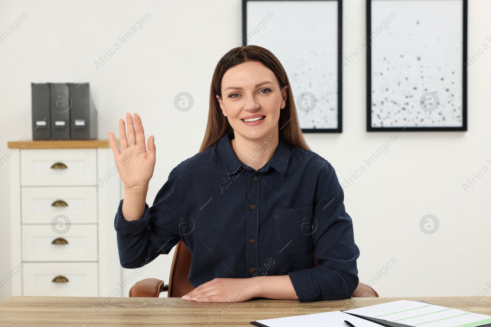 Photo of Woman waving hello during video chat at wooden table in office, view from web camera