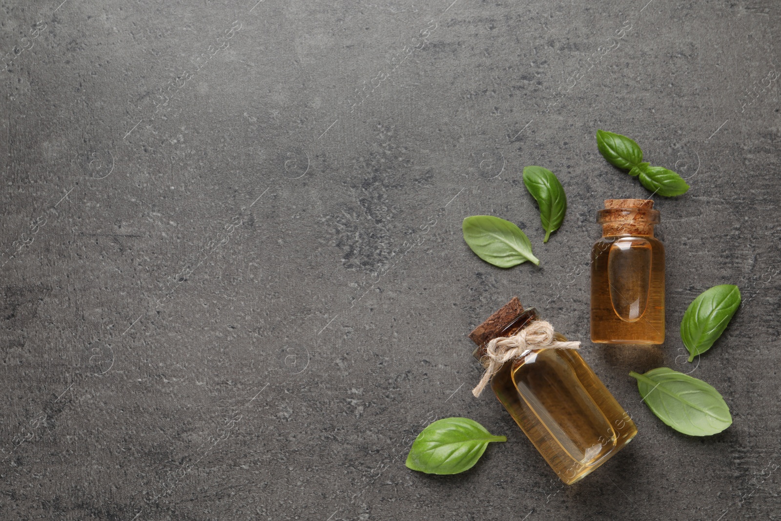 Photo of Bottles of essential basil oil and fresh leaves on grey table, flat lay. Space for text