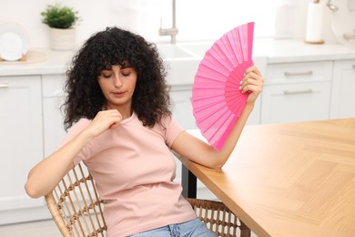 Photo of Young woman waving pink hand fan to cool herself at table in kitchen