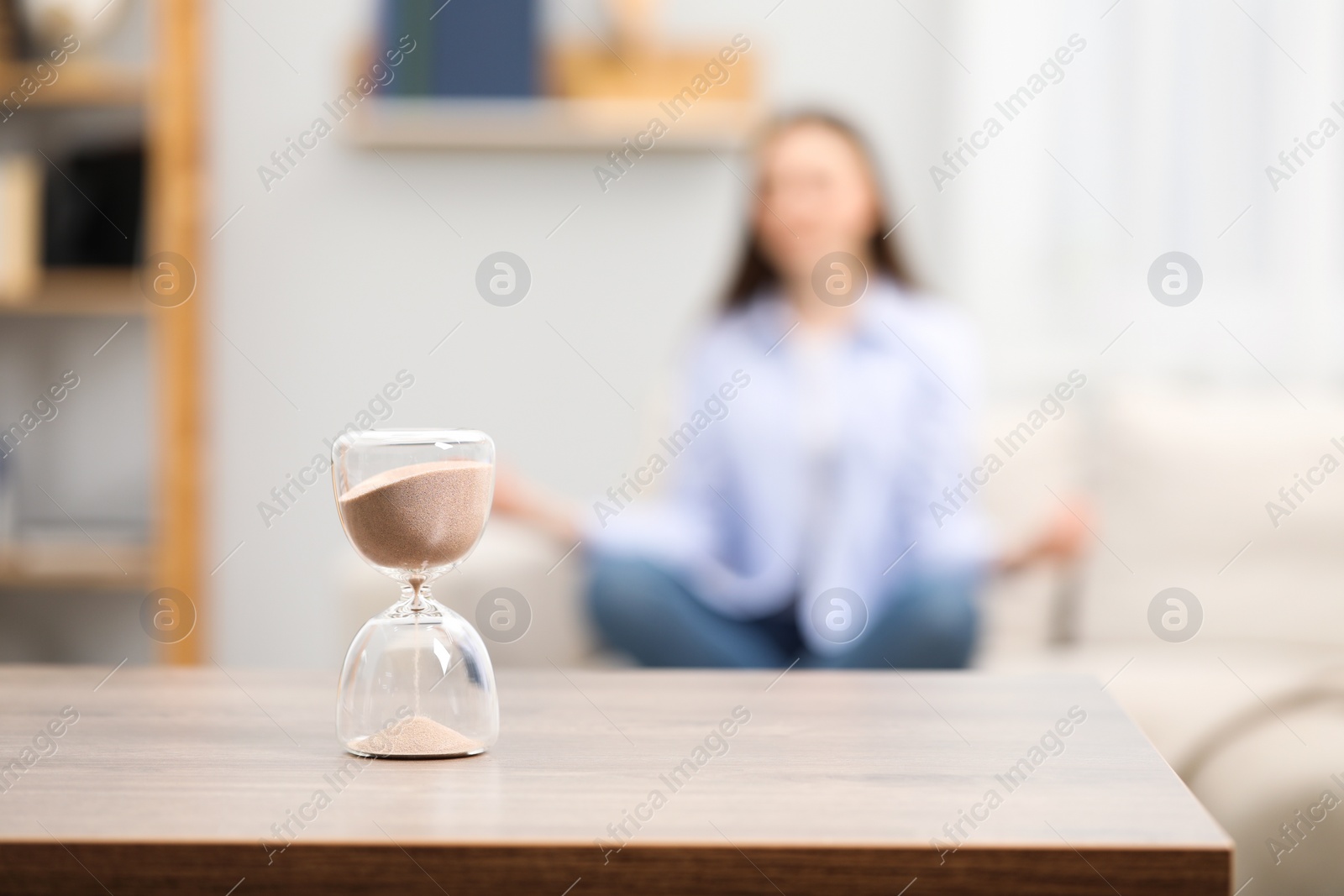 Photo of Hourglass with flowing sand on desk. Woman meditating indoors, selective focus