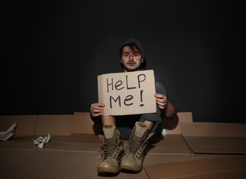 Photo of Poor young man with HELP ME sign on floor near dark wall