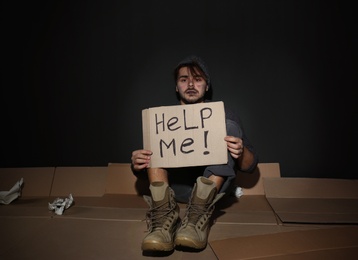 Poor young man with HELP ME sign on floor near dark wall