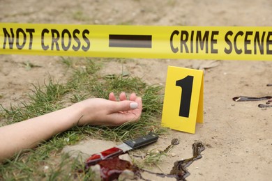 Crime scene with dead woman's body, marker, bloody knife and yellow tape outdoors, closeup