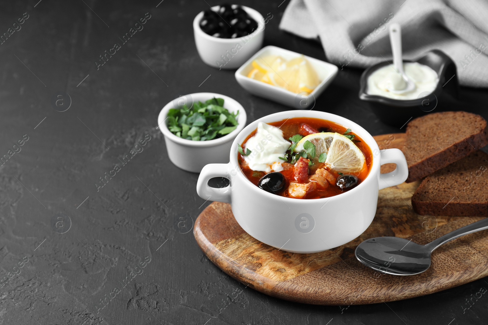 Photo of Meat solyanka soup with sausages, olives and vegetables in bowl served on dark grey textured table, space for text