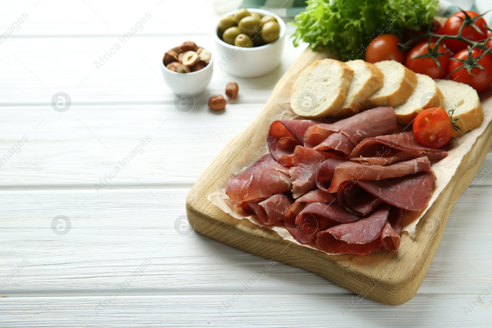 Photo of Board with delicious bresaola served with bread and tomato on white wooden table. Space for text