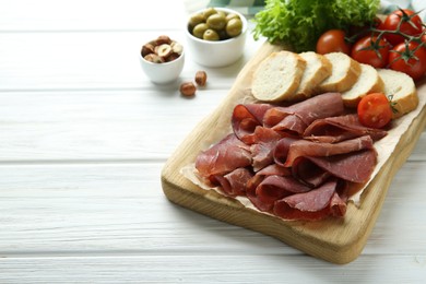 Board with delicious bresaola served with bread and tomato on white wooden table. Space for text