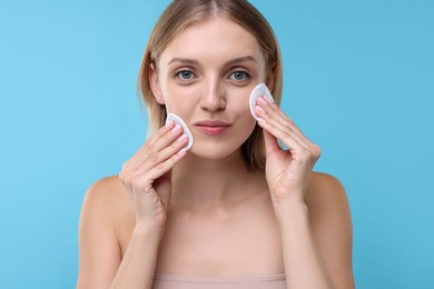 Young woman cleaning face with cotton pads on light blue background