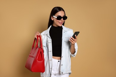 Young woman with stylish bag using smartphone on beige background