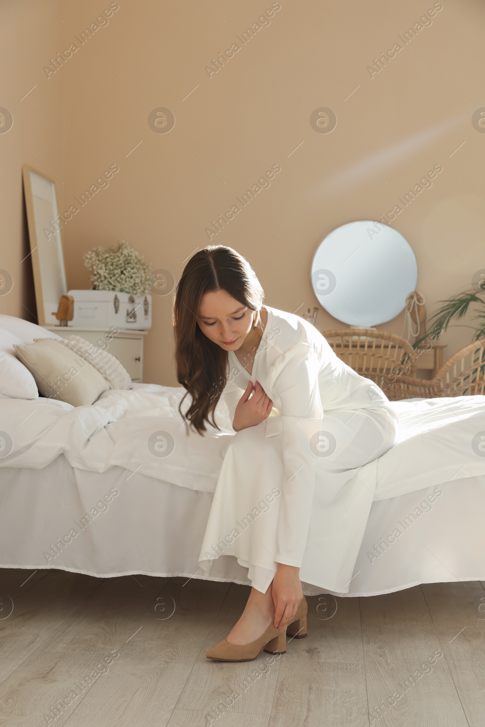 Photo of Beautiful young bride putting on stylish shoes in room. Wedding day