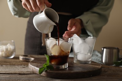 Photo of Woman pouring coffee into glass with ice cubes at wooden table, closeup
