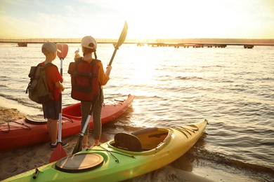 Photo of Children with paddles near kayaks on river shore, back view. Summer camp activity