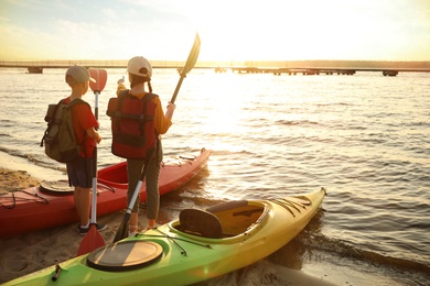 Children with paddles near kayaks on river shore, back view. Summer camp activity