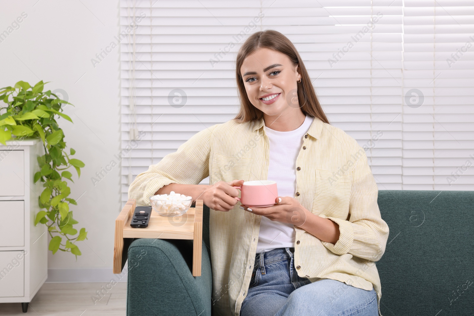 Photo of Happy woman holding cup of drink at home. Marshmallows and remote control on sofa armrest wooden table